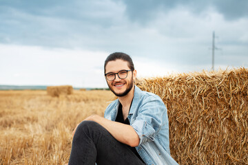 Naklejka na ściany i meble Portrait of young smiling guy sitting in wheat field near haystacks.