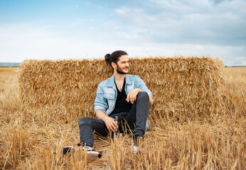 Naklejka na ściany i meble Young guy sitting on background of haystacks in wheat field.