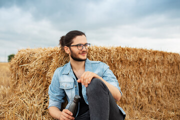 Naklejka na ściany i meble Young guy holding steel thermo bottle, sitting in field near haystacks.