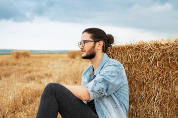 Side portrait of young guy in farm field sitting near haystacks.