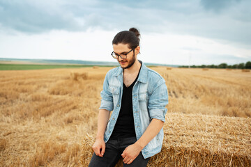 Naklejka na ściany i meble Confident smiling guy in denim jacket sitting on haystacks in farm field.