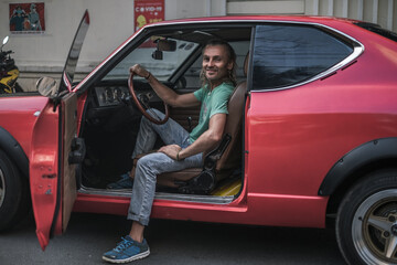 Mature handsome man sitting in red old style car, holding steering wheel and smiling. Long grey hair. 1970s style red car