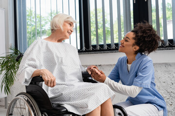 Obraz premium African american nurse holding hand of smiling elderly woman in wheelchair