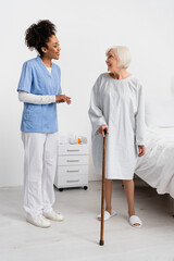 African american nurse looking at smiling senior patient with walking cane in hospital ward