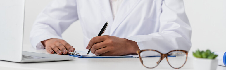 Cropped view of african american doctor writing on clipboard near laptop and eyeglasses in clinic, banner