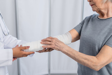 Obraz premium Cropped view of senior woman with plaster bandage on arm standing near african american doctor