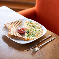 Eggs scrambled omelet with herbs toasts and tomato ketchup. Plate with food and cutlery on wooden restaurant table. Red armchair on blurred background. Soft focus. Copy space.