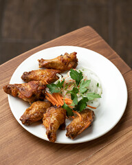 Fried chicken wings, onions, carrot and parsley on white platter. Roasted poultry meat. Food plate on wooden background. Vertical format. Soft focus.