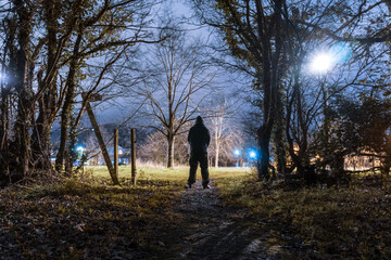 A hooded figure, silhouetted against a street light on the edge of town at night