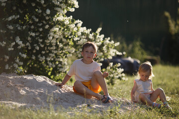 the older brother and his younger sister are sitting on the sand near a flowering jasmine bush, the boy is sad, and the girl is playing with the sand