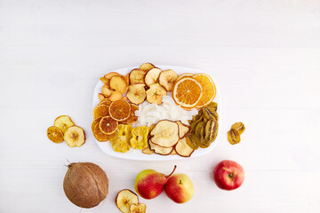 Dried apple, pear, pineapple,kiwi,apricot, coconut, orange and mandarin chips on plate on white wooden background. Vegan snack, sugar free food. 
