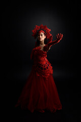 Fototapeta na wymiar Full length portrait of beautiful young asian woman wearing red corset, long opera gloves and ornate gothic queen crown. Graceful posing isolated on a dark studio background.