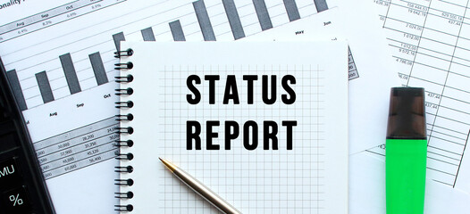 Text STATUS REPORT on the page of a notepad lying on financial charts on the office desk.