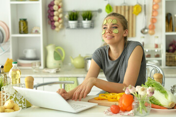 girl using laptop in kitchen with cucumber slices on face
