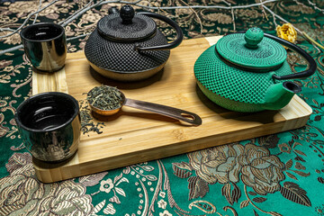 Asian tea service with two black and green cast iron teapots with copper saucepan with dehydrated tea inside and cups on fabric with gold embroidery