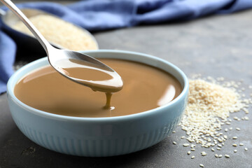 Spoon of tasty sesame paste above bowl on grey table, closeup