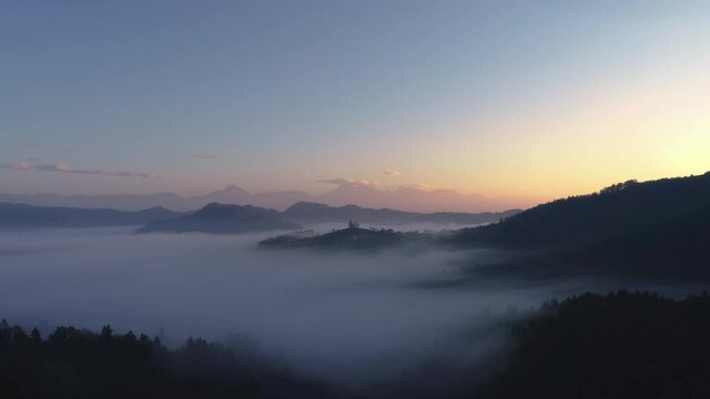 Aerial landscape of mountain hills covered in fog and Saint Thomas church, Slovenia on sunrise