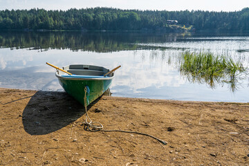 A green wooden rowing boat lies on the sandy shore of the reservoir. There is aquatic vegetation in...