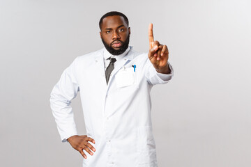 Serious-looking handsome african-american doctor in white coat, shaking index finger in warning,...