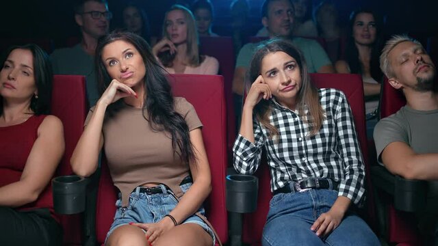 Two female spectators watching uninteresting film unpleasant face expression sleeping auditorium