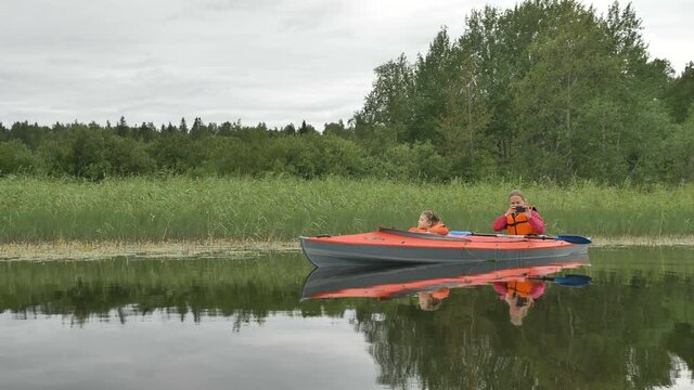 young blonde woman in vest takes picture with phone sailing reflecting in water kayak with daughter on lake through green reeds