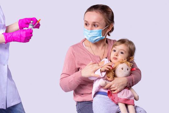 Caring Mother In Medical Mask Sitting On The Chair With Her Daughter On Her Knees Waiting For The Doctor To Make The Vaccine Against The Virus.