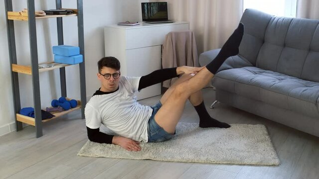 Young funny comical nerd man in glasses is doing fitness exercise for abs lying on carpet and moving legs at home. Training workout sport humor freak comic concept. Newcomer, beginner in sport.