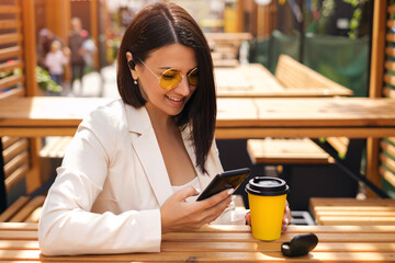A young beautiful woman is sitting at a table in a cafe on the terrace, drinking her coffee, writing messages in her smartphone and listening to music through wireless headphones. A business woman 
