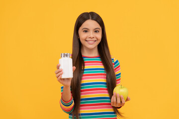 Happy girl child hold apple and supplement bottle yellow background, vitamins