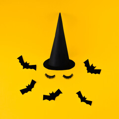 Creative witch look made of bats, hats and artificial beauty eyelashes. Minimum flat laying. Halloween concept. On a yellow background.