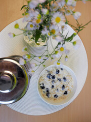 on a round plate with oatmeal porridge with coconut milk and blueberries, it lies on a table with a white round stand next to flowers and tea, next to white flowers . top view