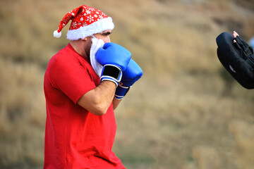 A man in a red sports T-shirt, Santa Claus hat and beard, and blue boxing gloves on his hands. He...