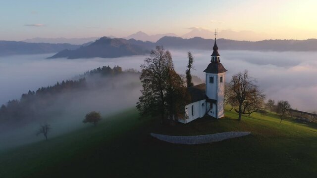 Aerial landscape of mountain hills covered in fog and Saint Thomas church, Slovenia on sunset