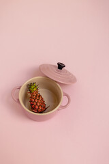 A mini pineapple in a small pink saucepan with a lid on a pink paper background with copy space. Creative food nutrients concept. Summer exotic fruit.