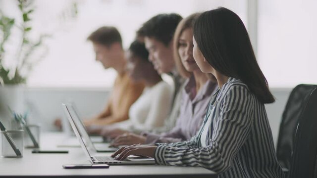 Group of diverse future IT specialists typing on laptop during lesson, asian girl asking for help groupmate, side view