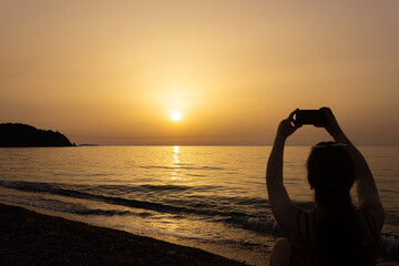 Silhouette of a person on the beach taking photograph with her mobile phone at dawn. Sun rises over the Mediterranean Sea on a lovely summer morning. Selective focus.