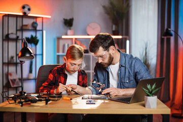 Obraz premium Caucasian boy and his father in protective glasses soldering together broken details from laptop. Man teaching his son how to repairing various modern technology.