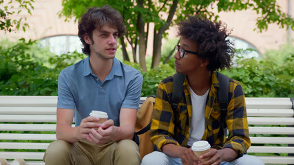 Multiethnic college students sitting on bench relaxing after classes drinking coffee