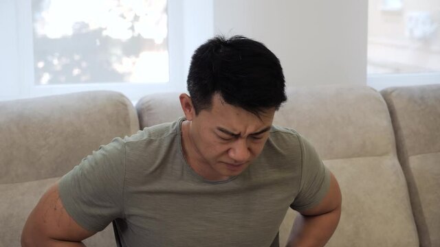 Asian Man feeling stomach pain at home, gastritis symptom, peptic ulcer, pancreatitis. Terrible pain in the stomach. A young man suffering from abdominal pain holding his stomach, sitting on the sofa.