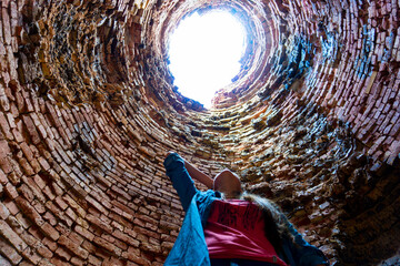 Deep stone well, light at the end of the tunnel. A girl stands at the bottom of an old blast furnace and looks up. Temperature damaged brick walls.