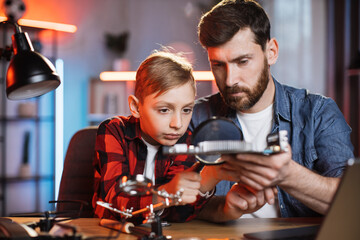 Bearded man and little boy using magnifying glass for examining problem of non working display card. Happy father with son trying to repair details from modern laptop.