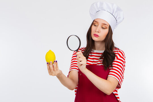 Woman in a chef's cap holds a lemon. She examines a lemon under a magnifying glass. Concept - chef selects citrus. Chef chooses a lemon for cooking. Girl in a red apron and a chef's hat - Powered by Adobe
