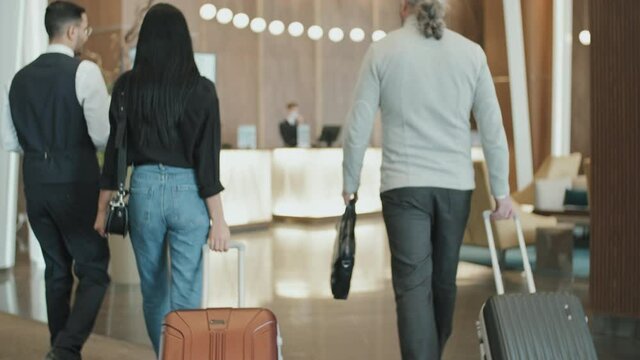 Tilting rear-view slowmo shot of young male concierge communicating with guest couple carrying luggage while escorting them to reception desk of posh hotel