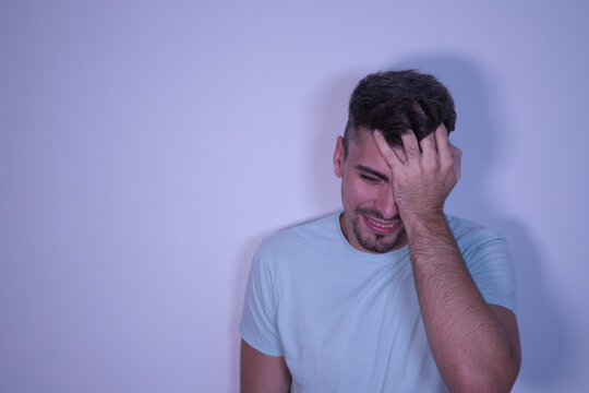 Young Hispanic male with hand on head and wincing in pain on white background.