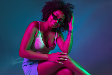 Portrait of African young girl posing isolated on dark blue studio background in pink neon light. Concept of human emotions, facial expression, youth, sales, ad.