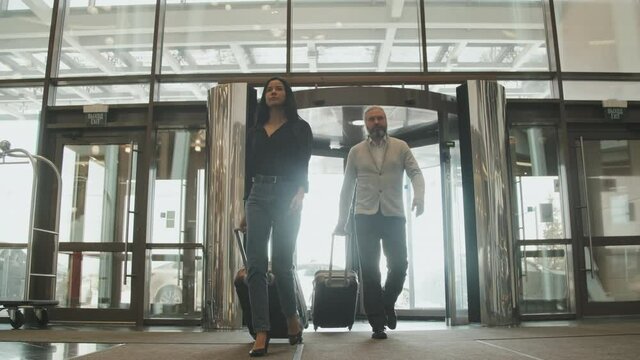 Low-angle full slowmo shot of modern couple with luggage walking through revolving door entering posh hotel for check-in