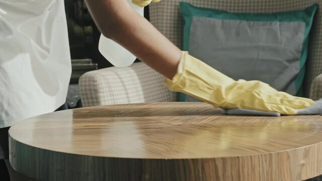 Slowmo close-up of housekeeper hands in rubber gloves cleaning and polishing surface of wooden coffee table in luxury hotel room