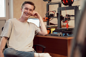 Cheerful young man sitting near 3D printers and laptop