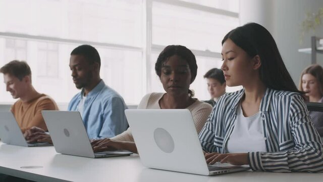Young asian girl having troubles during lesson, asking her black groupmate for help, typing on laptops at college
