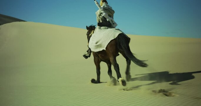 Masked person rides a galloping horse through the desert up a sand dune. Sand dunes surround a person riding a horse with a rifle on their back. Long journey. Cinematic, Shot on RED
